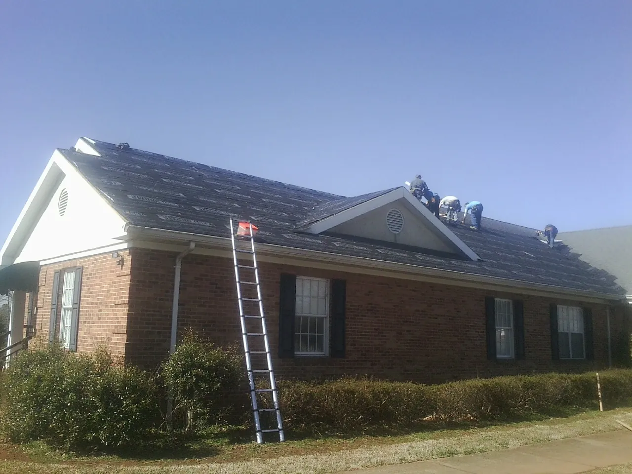 Crew tearing shingles off a brick ranch roof with a ladder set — Emergency Roof Repair work by our crew in Norman